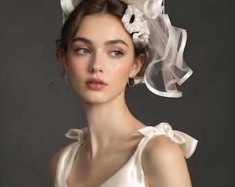 Puede incluir: Un vestido de satén color marfil con cuerpo sin mangas, dos lazos centrales, escote corazón y tirantes. El vestido se combina con un tocado a juego de organza transparente adornado con bucles de tela con volantes y detalles florales.
