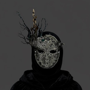 May include: A silver filigree masquerade mask with an elaborate, ornate design. The mask features a decorative headpiece with dark branches and a gold accent. The person is wearing a black hooded cloak, creating a mysterious and elegant look.