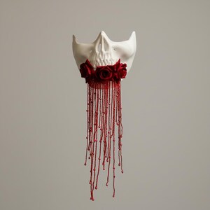 May include: A white skull mask adorned with red roses and flowing red beaded strands. The mask's skeletal features are prominent, with a neutral backdrop. The beaded strands create a dramatic effect.