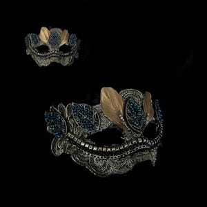 May include: An ornate silver-toned masquerade mask with blue and gold accents. The mask features intricate filigree detailing, blue bead embellishments, and gold-colored feather accents. The design is symmetrical and elegant, perfect for a formal event or costume.