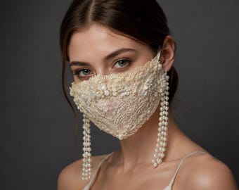 Puede incluir: Una mascarilla de color marfil confeccionada con tela de encaje intrincada, con delicados bordados florales, pequeños adornos de perlas blancas y dos largas hebras de perlas blancas que cuelgan de los lados de la mascarilla.