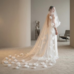 Puede incluir: Un velo de novia blanco con delicados adornos florales. El velo está decorado con flores en 3D y se extiende por el suelo. El vestido de novia es visible debajo del velo, creando un aspecto romántico. La longitud del velo se extiende por el suelo.