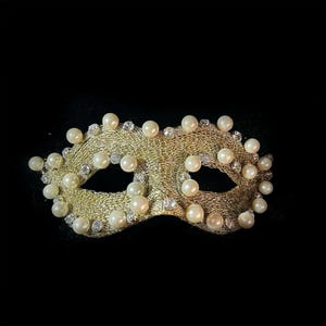 May include: A gold-colored masquerade mask with a mesh design, adorned with numerous faux pearls and small, sparkling rhinestones. The mask has eye openings and is set against a black background, creating a dramatic effect.