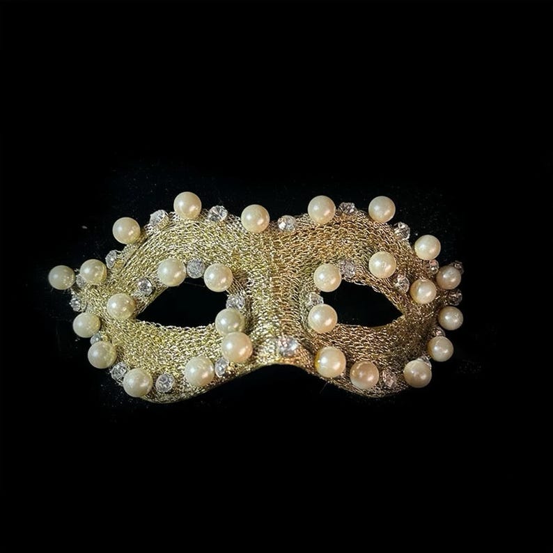 May include: A gold-colored masquerade mask with a mesh design, adorned with numerous faux pearls and small, sparkling rhinestones. The mask has eye openings and is set against a black background, creating a dramatic effect.