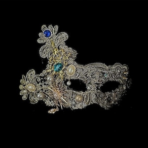 May include: An ornate, gold-colored masquerade mask with intricate filigree detailing. The mask is embellished with blue and green gemstones, pearls, and delicate floral accents. The mask is set against a black background.