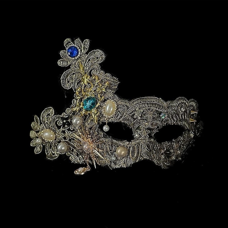 May include: An ornate, gold-colored masquerade mask with intricate filigree detailing. The mask is embellished with blue and green gemstones, pearls, and delicate floral accents. The mask is set against a black background.