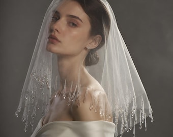 Puede incluir: Un velo de novia blanco y transparente de tul delicado, adornado con cuentas de cristal transparentes que cuelgan a lo largo del borde inferior. El velo está drapeado sobre la cabeza, mostrando el intrincado fleco de cuentas que atrapa la luz.