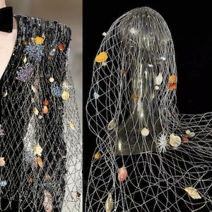 Puede incluir: Una prenda de terciopelo negro con una superposición de red decorativa, adornada con adornos coloridos en forma de concha. La red cubre los hombros y está adornada con varias formas y colores, creando un diseño artístico único.