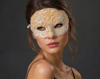 Puede incluir: Una máscara de baile de máscaras beige con intrincados bordados de encaje, patrones florales y pequeñas cuentas blancas. La máscara cubre el área de los ojos y está diseñada con bordes festoneados, mostrando un trabajo detallado.