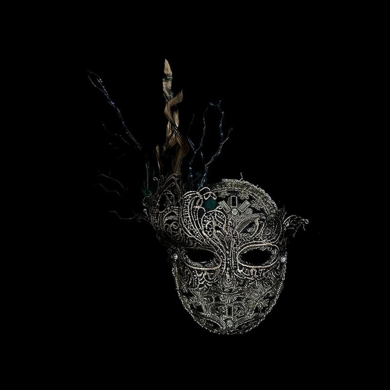 May include: Ornate silver masquerade mask with intricate lace-like detailing and embellishments. The mask features a decorative crown of branches and a dark background, creating a dramatic effect. Perfect for a masquerade ball or costume party.