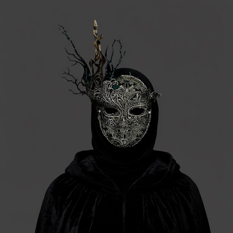 May include: A silver filigree masquerade mask with an elaborate, ornate design. The mask features a decorative headpiece with dark branches and a gold accent. The person is wearing a black hooded cloak, creating a mysterious and elegant look.
