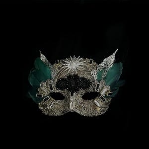 May include: A gold and silver masquerade mask with intricate lace detailing, adorned with black and clear gemstones, and accented with green feathers. The mask has a starburst design and is set against a black background.