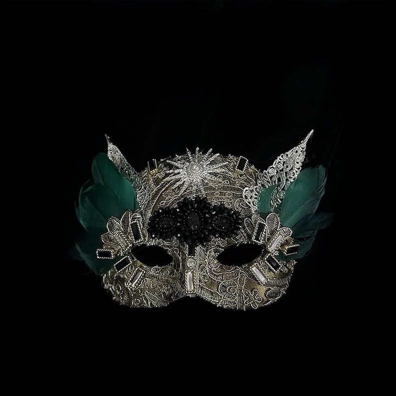 May include: A gold and silver masquerade mask with intricate lace detailing, adorned with black and clear gemstones, and accented with green feathers. The mask has a starburst design and is set against a black background.