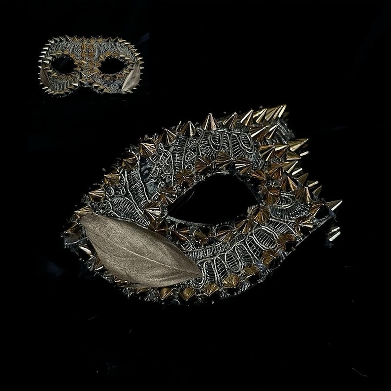 May include: A decorative eye mask with a detailed, ornate design. The mask is primarily silver and black, with gold-colored spikes and accents. A smaller mask is visible in the background. The mask has a leaf-shaped embellishment.