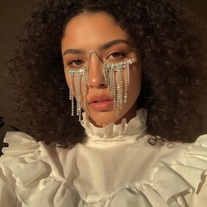Puede incluir: Primer plano de una persona que lleva un accesorio facial plateado con adornos de cristal colgantes. El accesorio enmarca los ojos y cae en cascada por la cara. La persona lleva una blusa blanca con volantes.