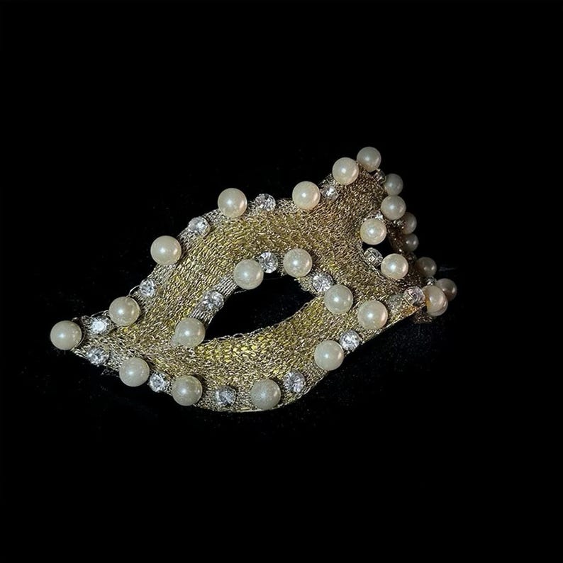 May include: A gold-colored masquerade mask with a woven design, adorned with faux pearls and clear rhinestones. The mask is set against a black background, highlighting its intricate details and elegant appearance.