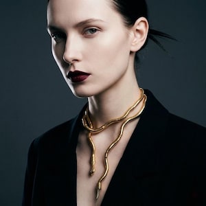 May include: A gold-toned snake necklace draped around the neck. The necklace has a detailed, textured design, with the snake's head and tail visible. The model is wearing a black blazer against a dark gray background.