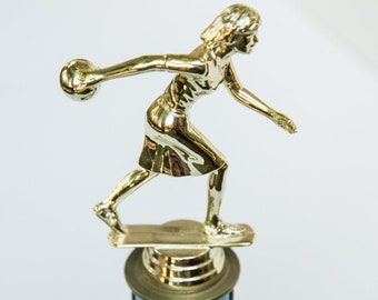 May include: A gold-colored trophy featuring a figure in a dynamic throwing pose, holding a small sphere. The figure stands on a cylindrical column with alternating light blue and gold vertical panels, mounted on a square dark brown wooden base.
