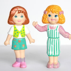 Peut inclure: Deux poupées vintage avec des traits peints. L'une a des cheveux bruns, une chemise blanche, un gilet vert à pois jaunes et une jupe turquoise avec des motifs de fleurs oranges. L'autre a des cheveux blonds bouclés, une robe rayée rose et des chaussures roses.