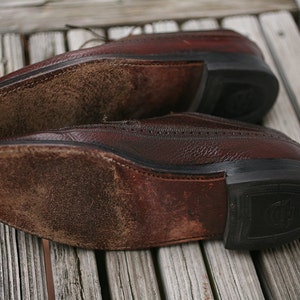 May include: Close-up of the soles of two brown leather shoes. The shoes have a worn, distressed look and are made of a textured leather. The soles are made of a black rubber material.