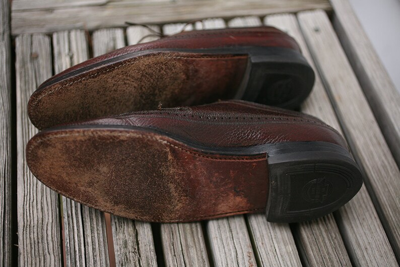 May include: Close-up of the soles of two brown leather shoes. The shoes have a worn, distressed look and are made of a textured leather. The soles are made of a black rubber material.