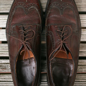 May include: A pair of brown leather shoes with wingtip detailing and laces. The shoes have a textured leather upper and a smooth leather sole.