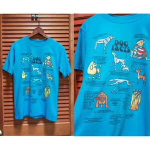 May include: A turquoise blue t-shirt featuring a graphic print of various dog breeds and facts about dogs. The text on the shirt reads "Dog Facts".