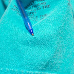 May include: A close-up of a turquoise blue t-shirt with a small hole in the fabric. A blue pen is lying on top of the shirt.