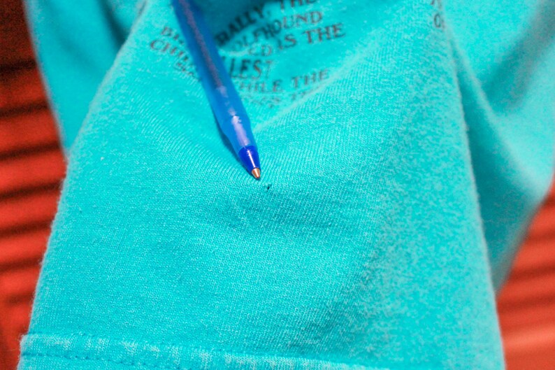 May include: A close-up of a turquoise blue t-shirt with a small hole in the fabric. A blue pen is lying on top of the shirt.