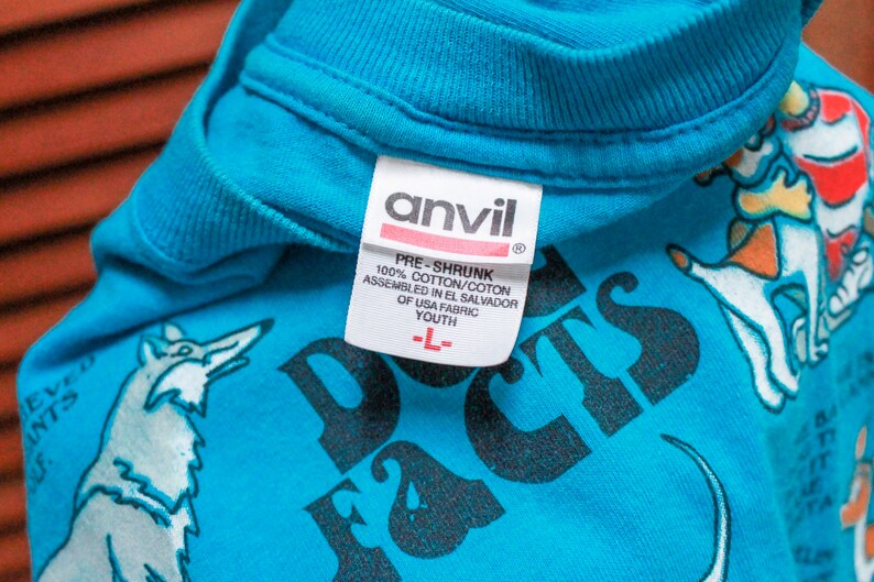 May include: A turquoise blue t-shirt with a black and white graphic print of dogs and the text "Dog Facts". The shirt has a white tag with the text "Anvil" and details about the fabric and size.
