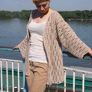 Op de afbeelding: Een beige gehaakt vest met een losse pasvorm en open voorkant. Het vest is gemaakt met een delicaat steekpatroon en heeft lange mouwen.