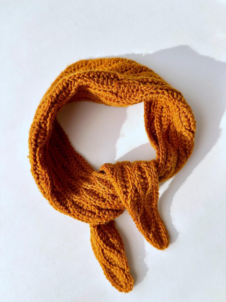 May include: Mustard yellow knitted scarf with a textured pattern. The scarf is tied in a knot, with the ends hanging down. The scarf is made of a soft, warm material, perfect for cold weather. The scarf is a versatile accessory.