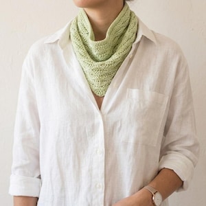 May include: A pale green knitted cowl scarf is worn over a white button-down shirt. The cowl is a soft, textured knit, and the shirt has a relaxed fit. The watch on the wrist has a gold band and a round face. The background is a plain, off-white wall.