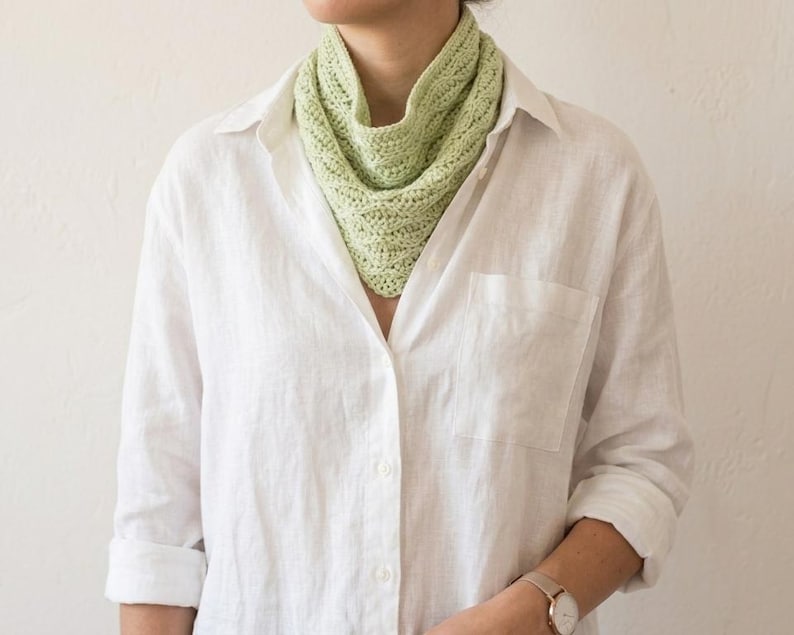 May include: A pale green knitted cowl scarf is worn over a white button-down shirt. The cowl is a soft, textured knit, and the shirt has a relaxed fit. The watch on the wrist has a gold band and a round face. The background is a plain, off-white wall.
