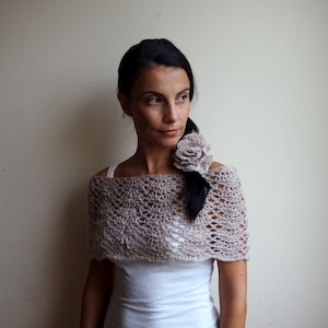 Op de afbeelding: Een vrouw draagt een beige gehaakte sjaal met een gehaakt bloemenaccent. De sjaal is een delicaat, opengewerkt ontwerp.