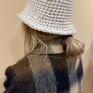 May include: A light beige crocheted bucket hat with a slightly slouched crown. The hat is worn by a person with long blonde hair pulled back in a ponytail. The person is wearing a brown, black, and white plaid fuzzy jacket.
