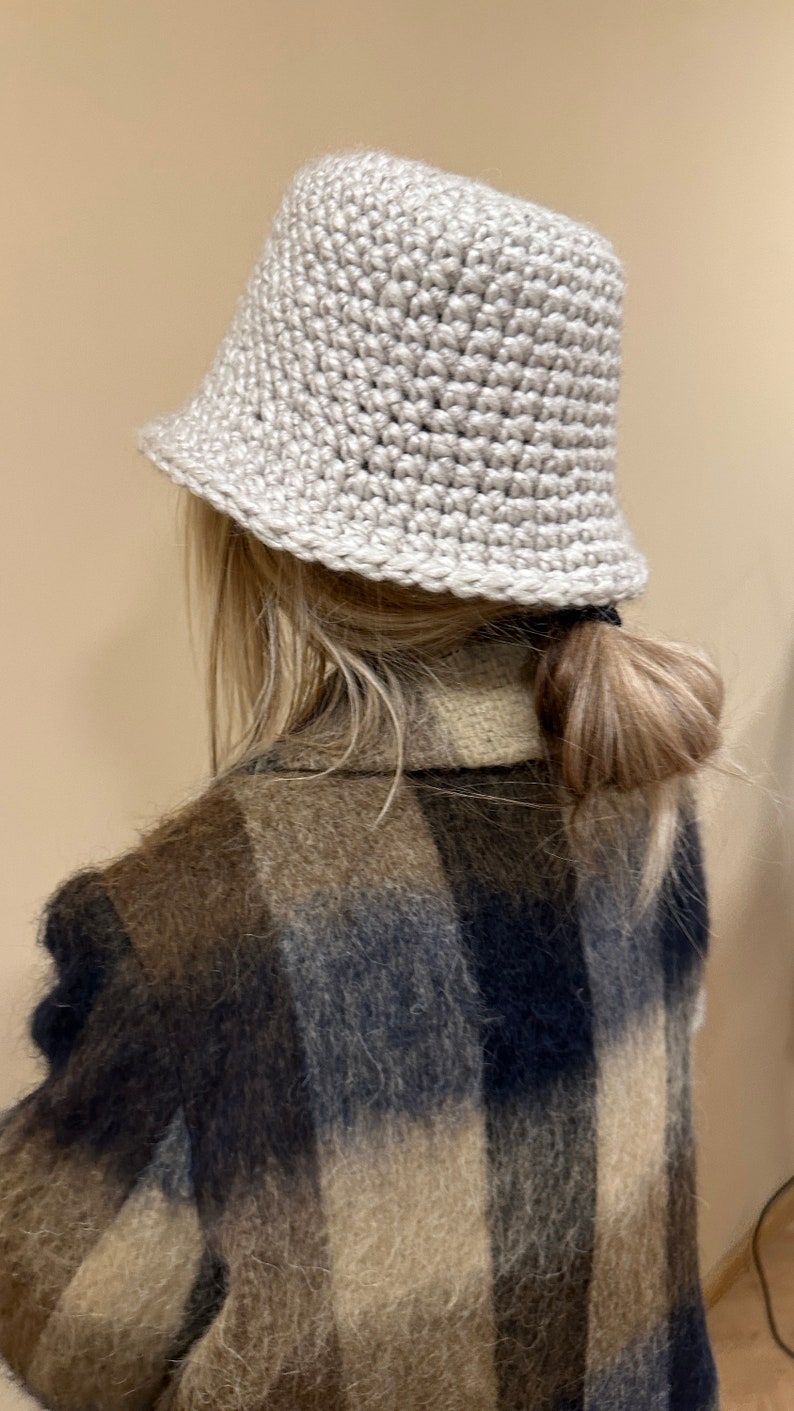 May include: A light beige crocheted bucket hat with a slightly slouched crown. The hat is worn by a person with long blonde hair pulled back in a ponytail. The person is wearing a brown, black, and white plaid fuzzy jacket.