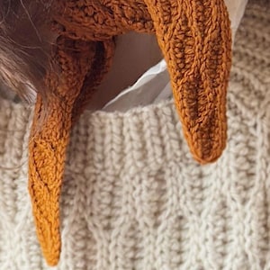May include: Close-up of a knitted scarf in a warm, burnt orange colour. The scarf is wrapped around a person's neck, with the ends hanging down. The background includes a cream-coloured, textured knit sweater.
