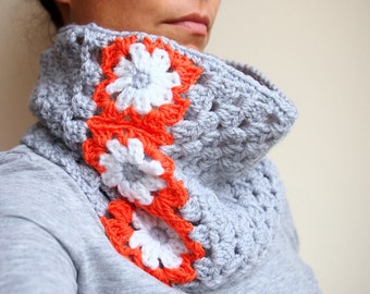 May include: A grey crocheted cowl with a white and orange floral design. The cowl is being worn by a person, but their face is not visible.