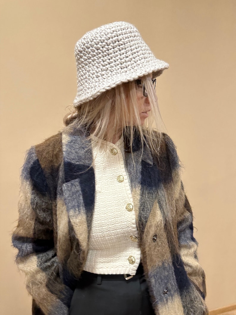 May include: A woman wearing a white crocheted bucket hat, a white sweater with gold buttons, a brown and blue plaid coat, and black pants.