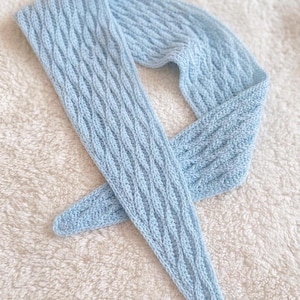 May include: A pale blue knitted scarf featuring a cable knit design. The scarf is made from soft yarn and is displayed on a white, textured surface. The scarf is long and narrow, suitable for wearing around the neck.