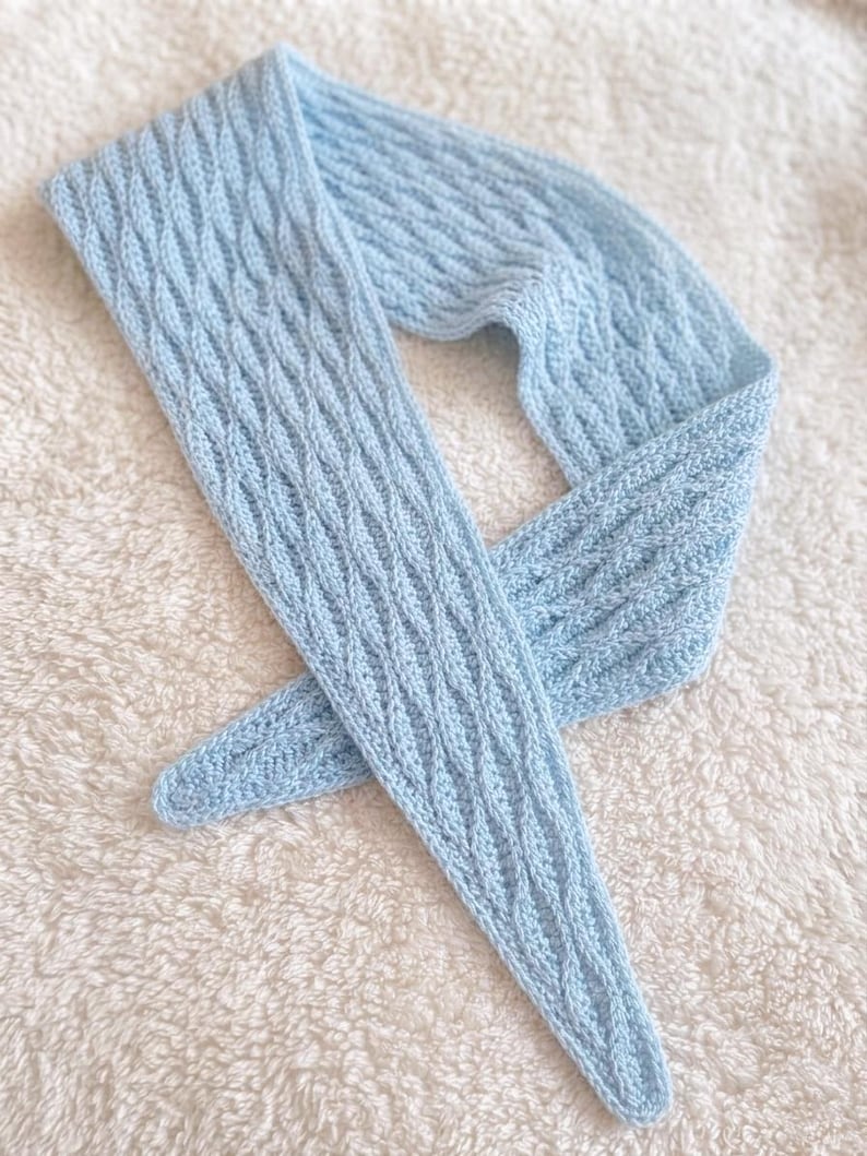 May include: A pale blue knitted scarf featuring a cable knit design. The scarf is made from soft yarn and is displayed on a white, textured surface. The scarf is long and narrow, suitable for wearing around the neck.