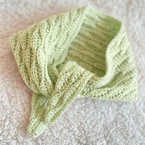 May include: A light green, handmade crochet cowl scarf. The scarf has a textured stitch pattern and is folded over on a white, textured surface. The cowl is a soft, pastel green colour.