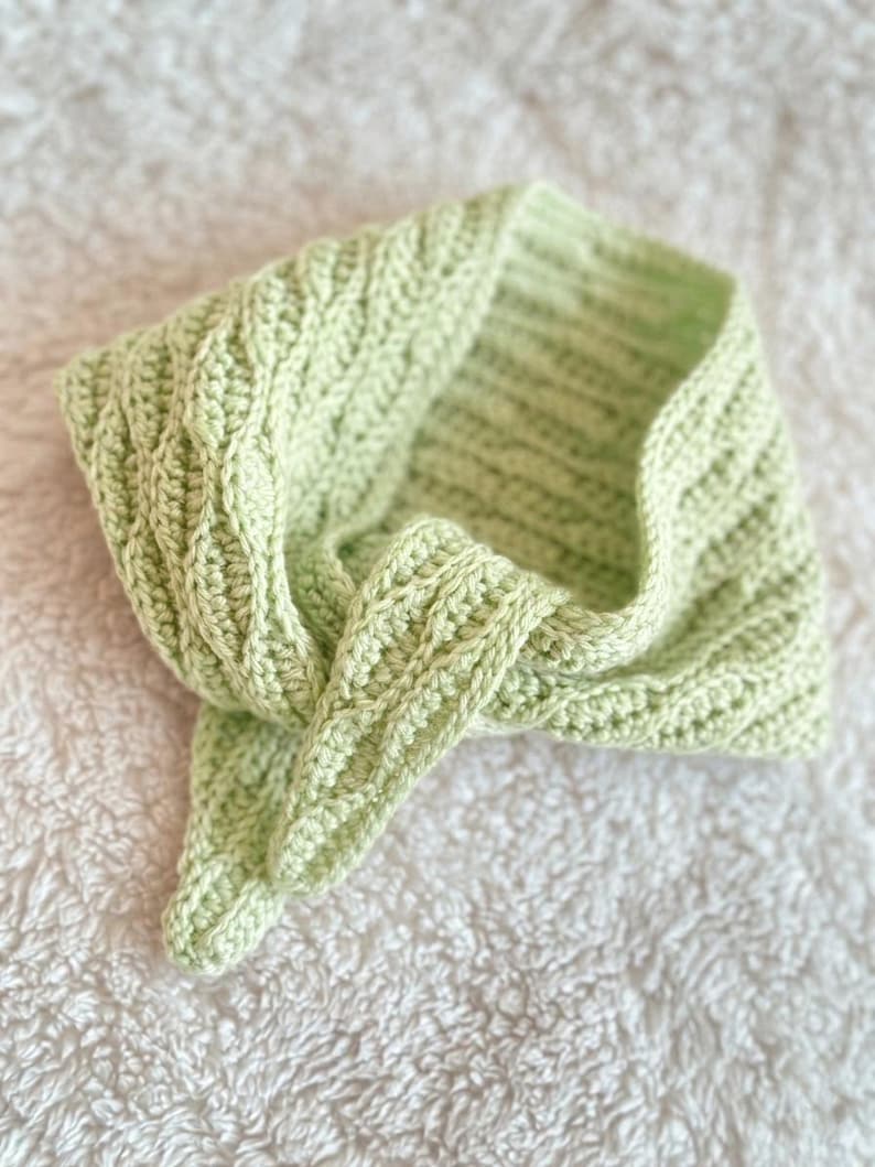 May include: A light green, handmade crochet cowl scarf. The scarf has a textured stitch pattern and is folded over on a white, textured surface. The cowl is a soft, pastel green colour.