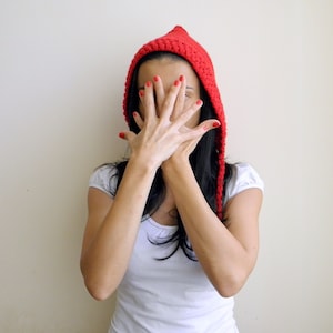 May include: A vibrant red, crocheted hood with long ties. The hood is worn over black hair and paired with a white t-shirt. The person is covering their face with their hands, with red nail polish.