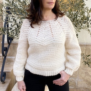 Puede incluir: Un suéter de crochet blanco con un delicado patrón de encaje alrededor del cuello y las mangas. El suéter tiene un ajuste holgado y una longitud ligeramente recortada.