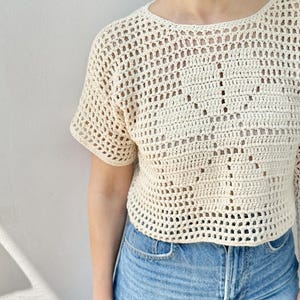 May include: Cream-colored crochet top with short sleeves. The open weave design creates a textured look. The top is paired with blue denim jeans. The top is a casual, handmade garment.