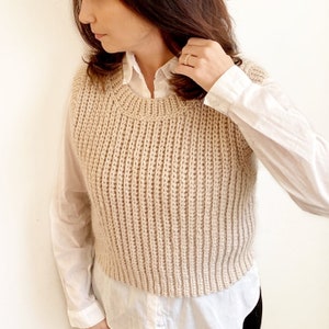以下が含まれることがあります： ベージュのリブ編みニットベスト。ノースリーブの衣服はクルーネックとクロップドフィットです。ベストは白い襟付きシャツの上に着用されています。リブ編みのパターンは垂直で、ベストの全長にわたって走っています。
