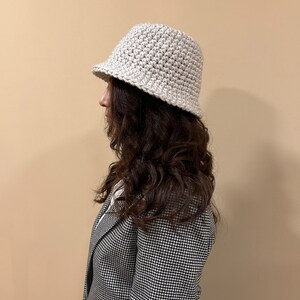 May include: A white crocheted bucket hat with a slightly slouched crown. The hat is worn by a person with long brown hair and a black and white houndstooth blazer.
