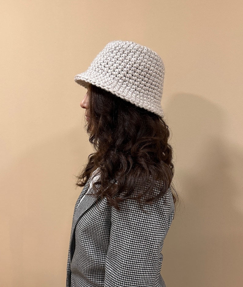 May include: A white crocheted bucket hat with a slightly slouched crown. The hat is worn by a person with long brown hair and a black and white houndstooth blazer.